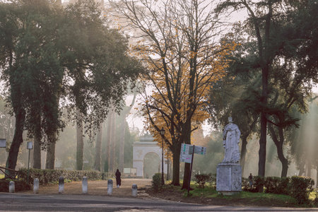 Serene scene of tranquil park surrounded by tall trees and prominent statue. Soft morning light...