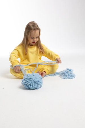 Pretty little girl in yellow clothes plays with ball of blue thread. Child model with long blond hair sits in studio on light background. High quality photoの写真素材
