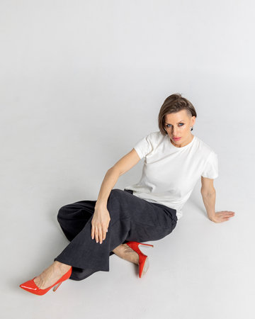 Portrait beautiful woman of model appearance. Girl in white T-shirt, black trousers, red shoes on grey background. Pretty female with trendy makeup posing in studio. Vertical photo.の写真素材