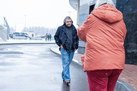 Female photographer in red jacket filming man in headphones listening to music in snowy rainy...