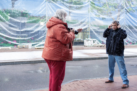 Female photographer in red jacket filming man in headphones listening to music in snowy rainy weather outside modern business center. High quality photoの写真素材