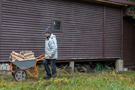 Adult man pushing wheelbarrow loaded with firewood along stone path in yard wearing warm jacket and hat.の写真素材