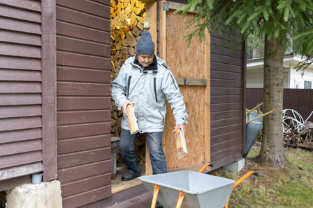 Man moving stacked logs in wheelbarrow near house and fence on rural property during cool season.の写真素材