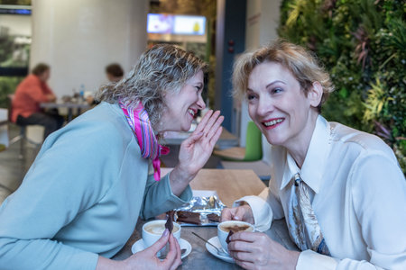 Two mature women sitting at wooden table in modern cafe holding cups of coffee and dessert engaged...