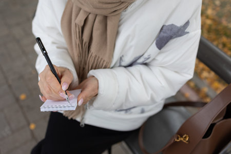 Thoughtful female in white jacket and beige scarf writing notes while standing by metal bench in...
