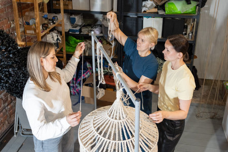 Teaching mentor assisting women during hands on masterclass creating large woven pendant lamps from metal frames and rope.の写真素材