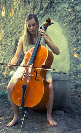 Beautiful young blonde girl with white dress playing a cello in a sunset scenarioの写真素材