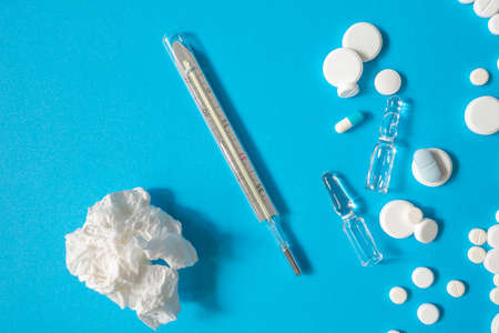 Thermometer, ampoules, pills and crumpled paper napkin on blue paper background. Concept of common cold, or flu. Copy spaceの写真素材