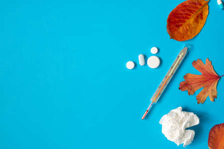 Thermometer, pills, crumpled paper napkin and yellow fallen leaves on blue paper background.