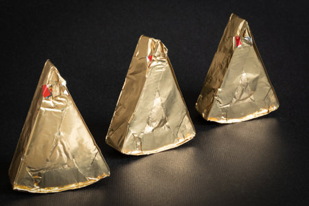 Thre triangular pieces of melted cream cheese wrapped in a golden aluminum foil on a black...