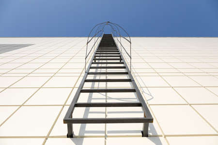 A long metal staircase on the gray modern facade of an industrial building, warehouse or shopping center against the blue sky. Symbol of the stairway to heaven. Fire fighting equipment conceptの写真素材