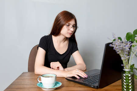An attractive young woman is sitting in front of a laptop, shopping on the Internet, chatting with friends or just surfing the web. A cup of coffee on the tableの写真素材