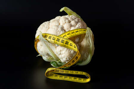A fresh cauliflower head wrapped with a measuring tape on a black background. The concept of healthy nutrition, weight loss, dieteticsの写真素材