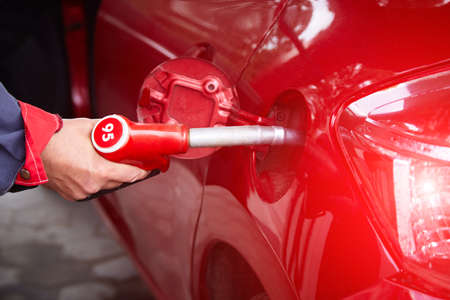 A man's hand holds a filling gun inserted into the hole of a gasoline tank of a car on a gasoline fueling. Close-up of the hand and fuel filling pistol. Red refueling gun at the refuel station.の写真素材