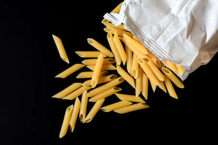Whole grain penne pasta spilled out of white paper bag on a black background. Pasta gourmet still life and pasta healthy eating. Top viewの写真素材