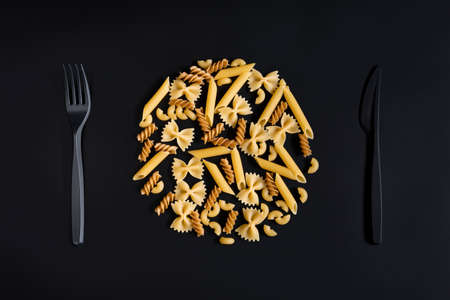 A variety of pasta in the shape of a plate and black plastic cutlery on a black background for menu design, a poster on a culinary theme. Allusion to the table settingの写真素材