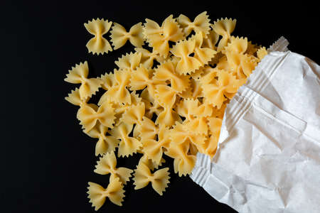 Whole grain farfalle pasta spilled out of white paper bag on a black background. Pasta gourmet still life and pasta healthy eating. Top viewの写真素材