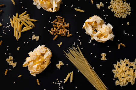 Different types of uncooked dried pasta on the black background. Raw pappardelle, penne, spaghetti, farfalle and fusilli. Italian pasta cooking conceptの写真素材