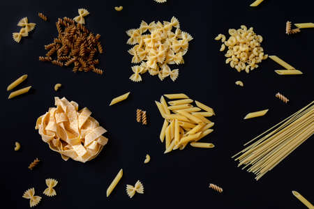Different types of uncooked dried pasta on the black background. Raw pappardelle, penne, spaghetti, farfalle and fusilli. Italian pasta cooking concept.の写真素材
