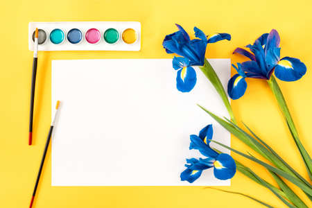 Three blue irises, a sheet of white paper, watercolor paints and brushes on a yellow background. copy spaceの写真素材