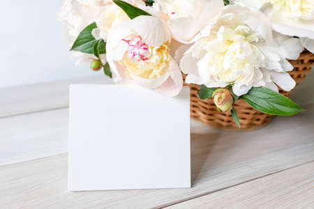 Beautiful fragrant peonies in a wicker basket and an empty greeting card on a white wooden table. Seasonal flowers as a gift for a birthday, wedding decoration or other celebration. Copy Spaceの写真素材