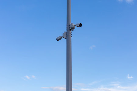 Two street surveillance cameras on a pole against a blue sky background. The cameras are facing in...