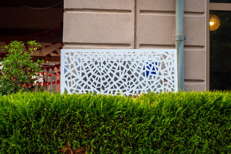 A decorative metal grate and trimmed bushes cover the air conditioning unit near the building wall....