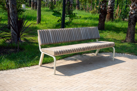 Outdoor seating in landscaped garden. Modern bench on stone walkway surrounded by grass and trees....