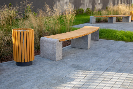Modern urban seating and trash bin in a landscaped area with grass and ornamental plants. City...