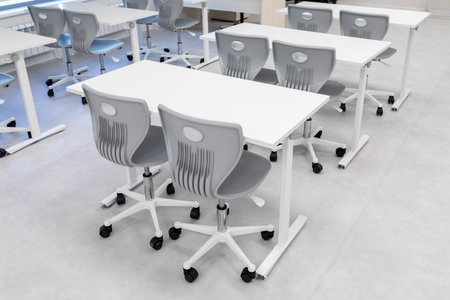 Modern classroom with white tables and gray rolling chairs on light floor. Educational furniture for...