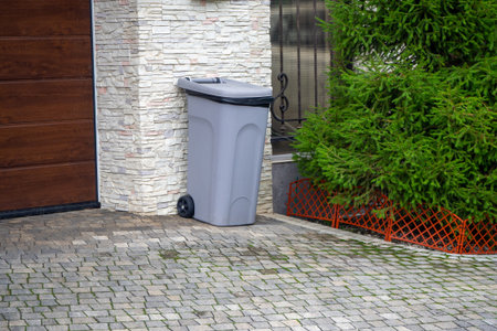 Gray plastic waste bin near garage wall on paved driveway surrounded by green plants. Waste management, urban maintenance, residential infrastructure, ecological design. Real photoの写真素材