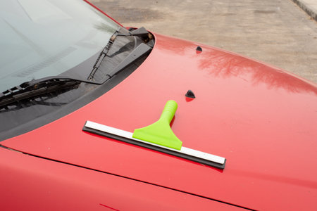 Green squeegee on red car hood near windshield. Car cleaning, auto maintenance, detailing tool. Real...
