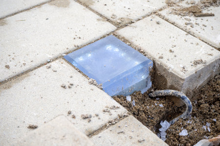 Transparent glass block embedded in paving stones with visible soil and wire. Landscape lighting, garden design, construction detail. Real photoの写真素材