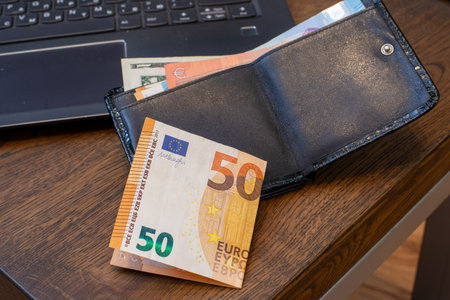 Black leather wallet with 50 Euro bill on wooden desk beside laptop. Finance, personal budget,...