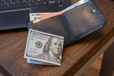 Black leather wallet with 100 US Dollar bill on wooden desk beside laptop. Finance, personal budget,...