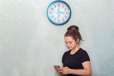 A young girl in headphones stands under the clock and looks at a mobile phone. On the wall clock, the hands seemed to have frozen in motion, time has stopped for the teenagerの写真素材