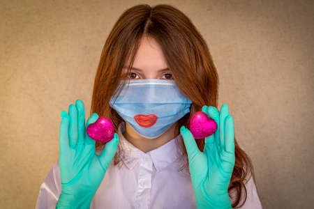A girl teenager wearing a medical mask and gloves, holds two scarlet hearts in front of her. Red lips are painted on the mask. Selective focusの写真素材