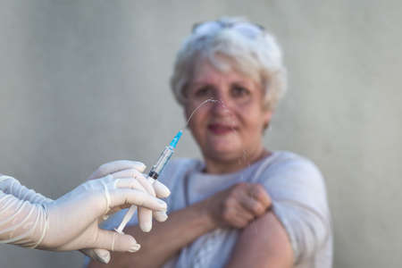 Hands in medical gloves are holding a syringe, preparing to vaccinate, in the background an elderly woman is smiling and ready to give the shot.の写真素材