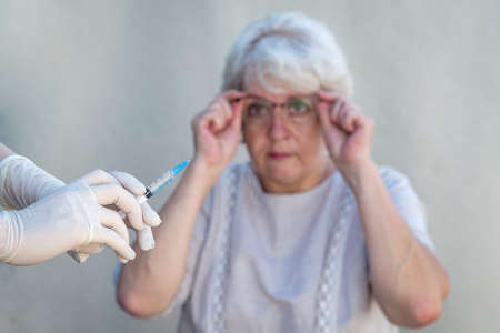 Hands in medical gloves holding a syringe, preparing for vaccination, in the background an elderly woman holding glasses, worried, unsure of her choiceの写真素材