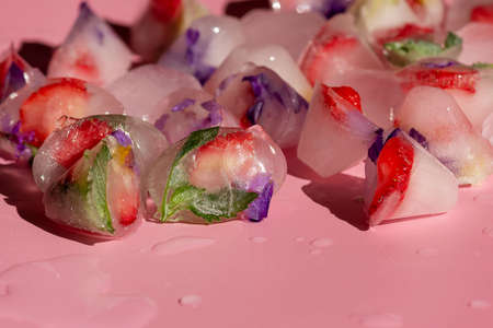Edible flowers, strawberry and mint in the form of ice cubes on a pink background, Close-up.Place...