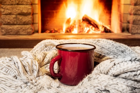 Red enameled mug for hot tea and cozy warm scarf near fireplace, in country house, winter vacation, horizontal.の写真素材