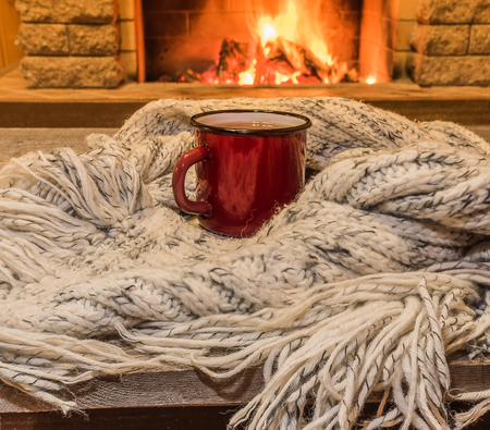 Red enameled mug for hot tea and cozy warm scarf near fireplace, in country house, winter vacation, horizontal.の写真素材