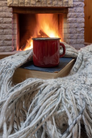 Red mug with tea, and warm wool scarf , a book , near cozy fireplace, in country house, winter vacation.の写真素材