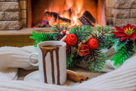 Big mug with hot chocolate, near cozy fireplace, christmas decoration and wool scarf, in country house, winter vacation.の写真素材
