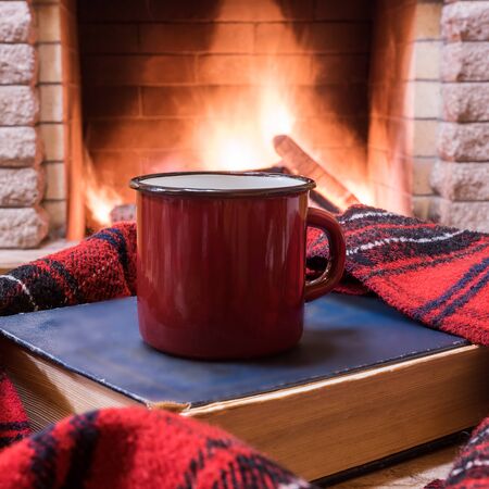 Mug of hot tea, wool scarf and book near cozy fireplace, in country house, winter vacation.の写真素材