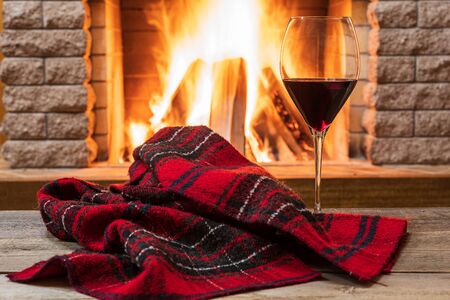 Glass of red wine and warm scarf against cozy fireplace background, in country house, horizontal, hygge , home sweet home.