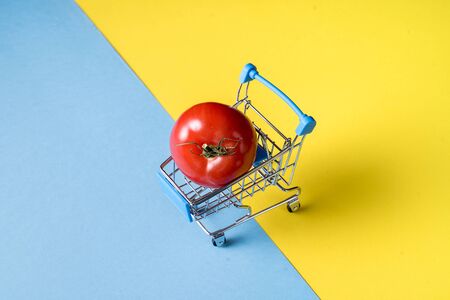 Empty shopping trolley carrying tomato on bright blue and yellow background. Copy space for text or design.の写真素材