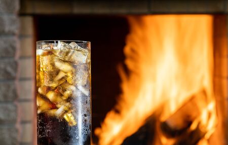 Glass of cola with ice in front of a burning fireplace in country house.の写真素材
