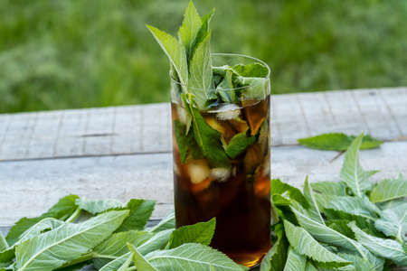 Cold iced tea with mint leaves and ice in tall glass on wooden table outdoor.