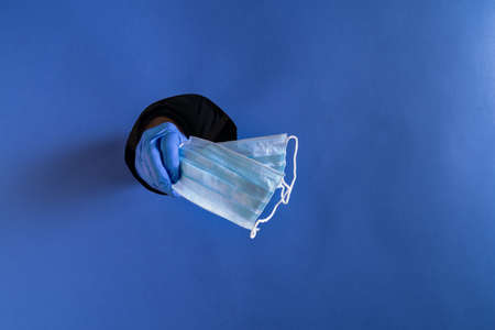 COVID-19 Concept. Coronavirus prevention medical face mask in hand through blue paper hole. Space...